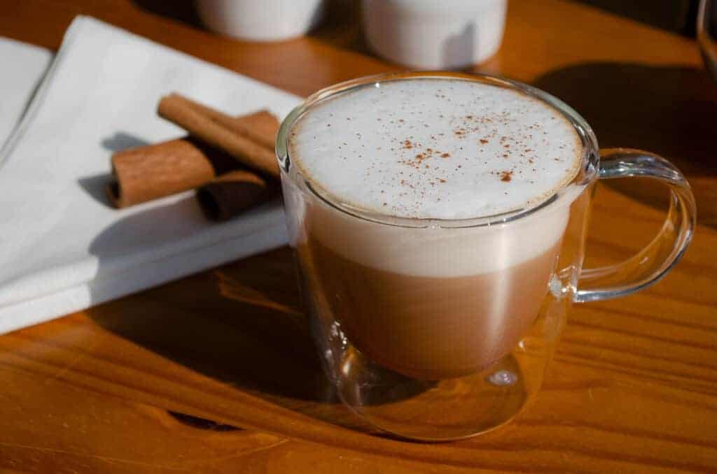 Receita de chápuccino em uma caneca de vidro duplo sobre a mesa. Ao lado, sobre um guardanapo, pedaços de canela.