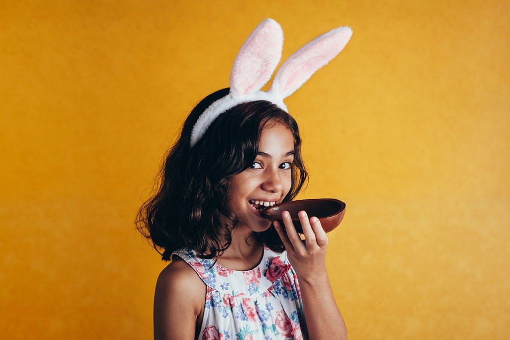 Foto de uma criança com orelhas de coelho decorativas, mordendo um ovo de páscoa de chocolate.
